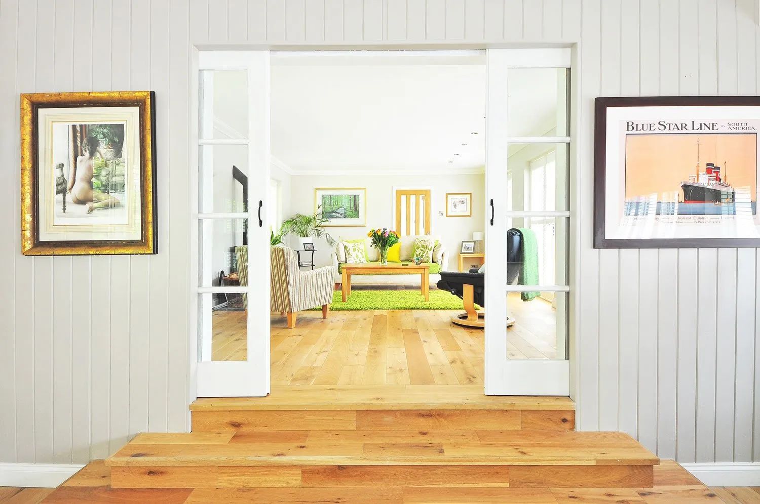 Open white French doors frame a bright living room with light wood floors, a green rug, and two framed wall artworks.