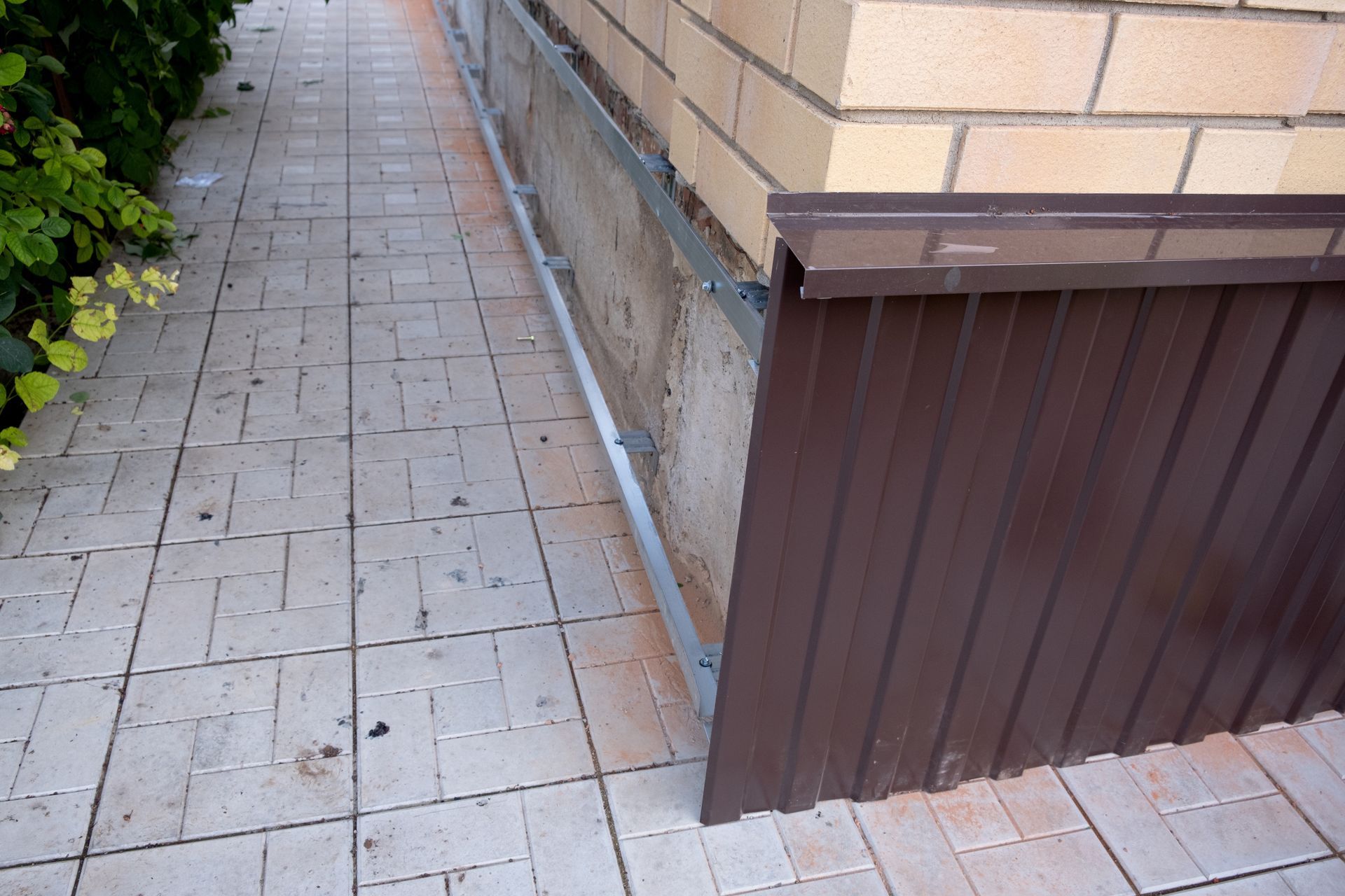 Narrow tiled sidewalk beside a beige wall and brown fence panel, with bushes lining the left side