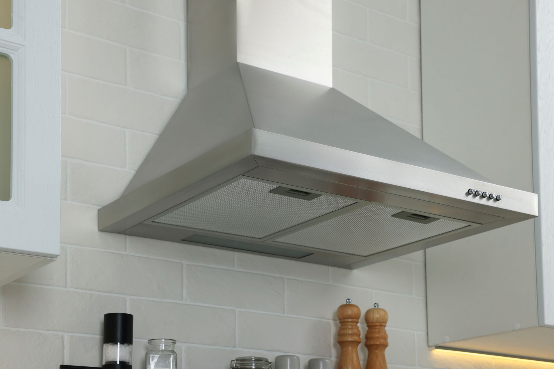 Stainless steel kitchen range hood above a stove, with white tile backsplash and cabinets.