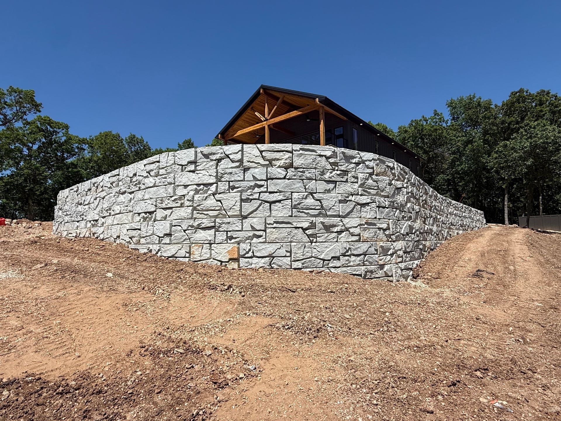 Stone retaining wall with a wooden pavilion on top, set on a dirt hillside under a clear blue sky