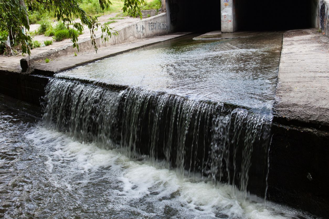 Water flowing over a small concrete weir into a canal, with greenery along the banks.