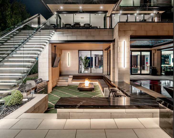Luxury outdoor living space with fire pit, grill, and tiered seating, at night.
