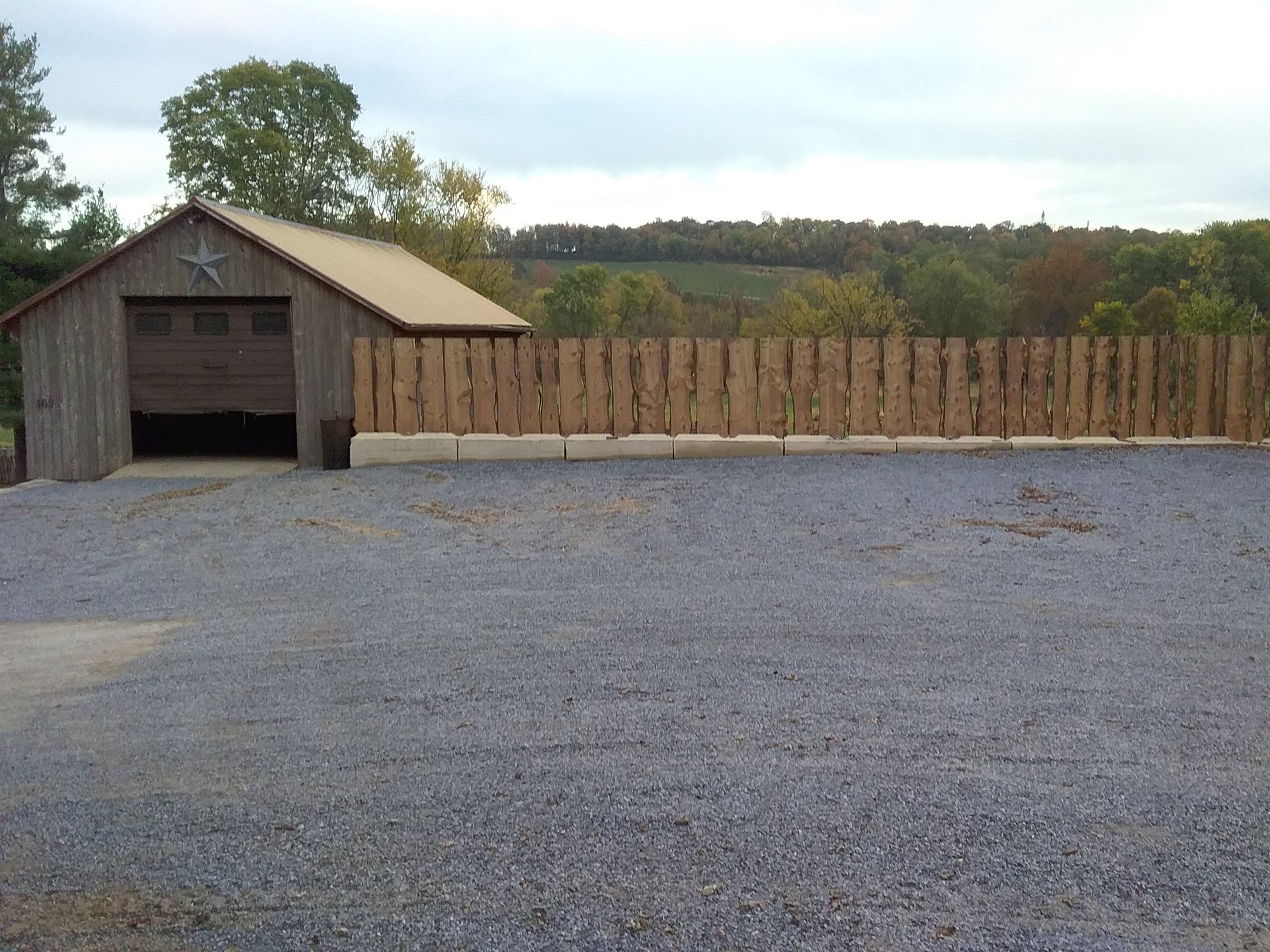 Small rustic shed beside a long wooden fence on a gravel lot, with trees and open field behind it