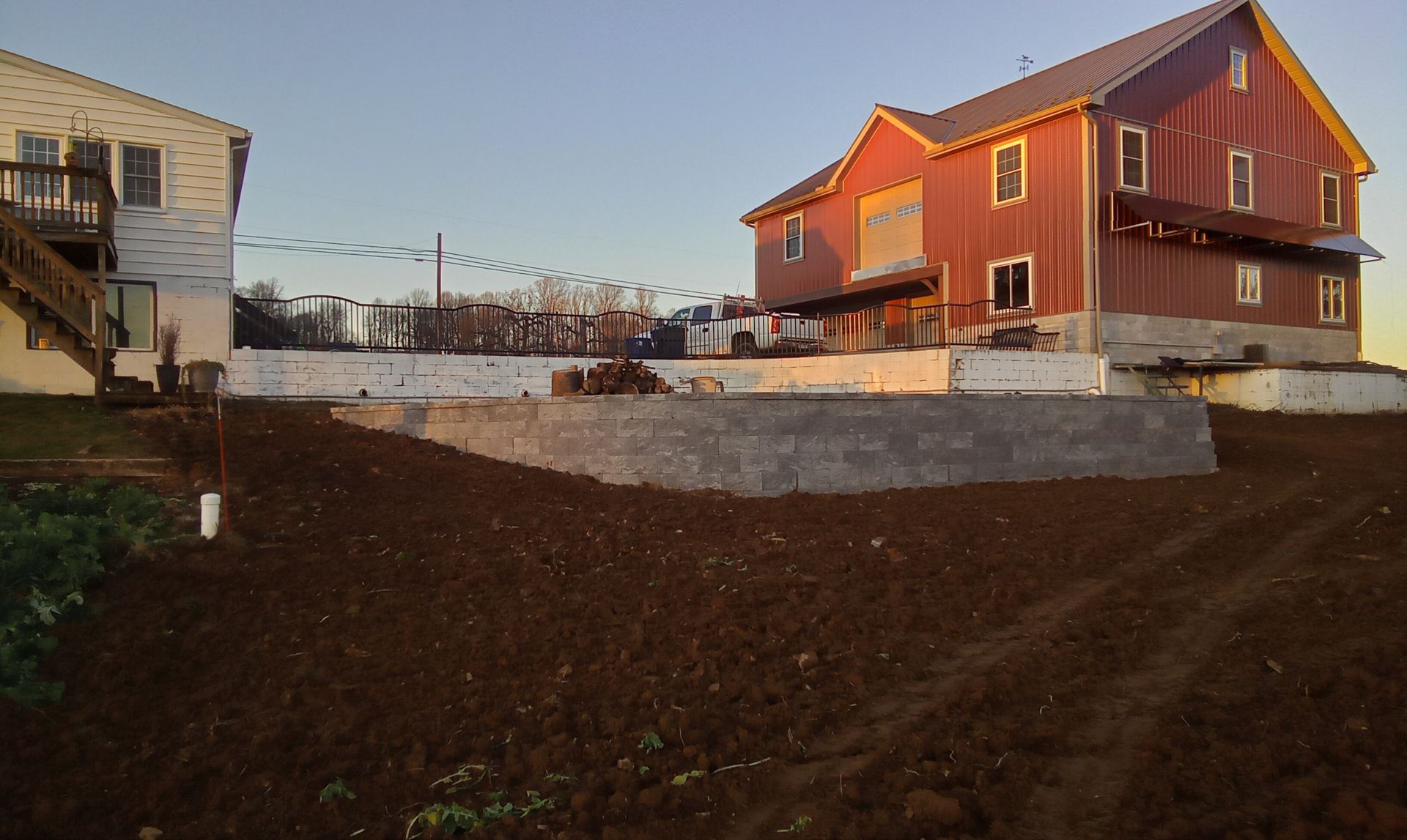 Red house beside a dug-up dirt lot at sunset, with neighboring homes and a low concrete wall.