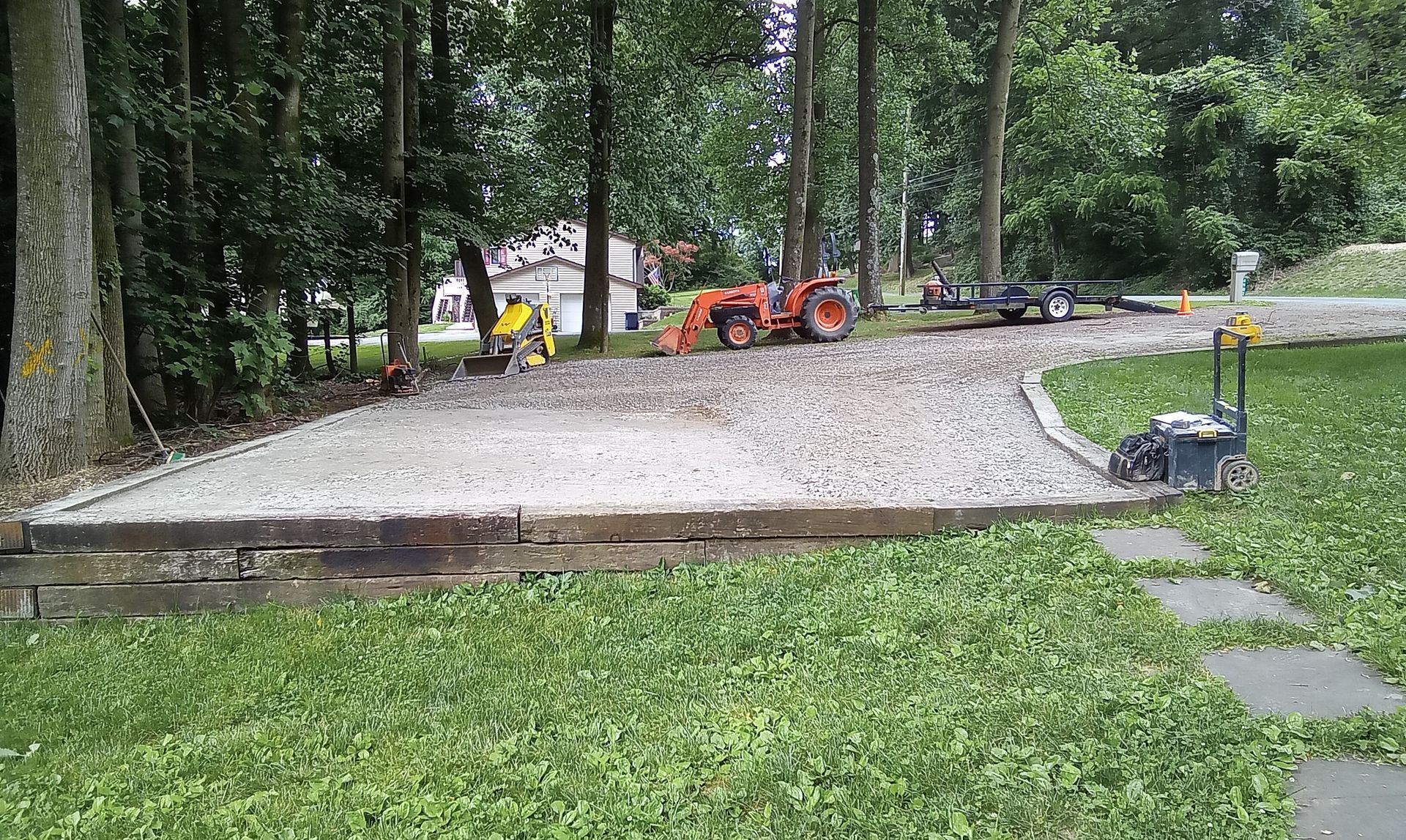 Gravel driveway construction with orange loader and workers near trees and lawn