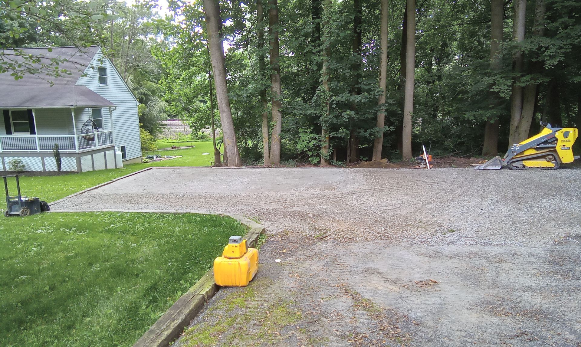Gravel driveway beside a wooded yard, with a yellow can in the foreground and a small house at left.