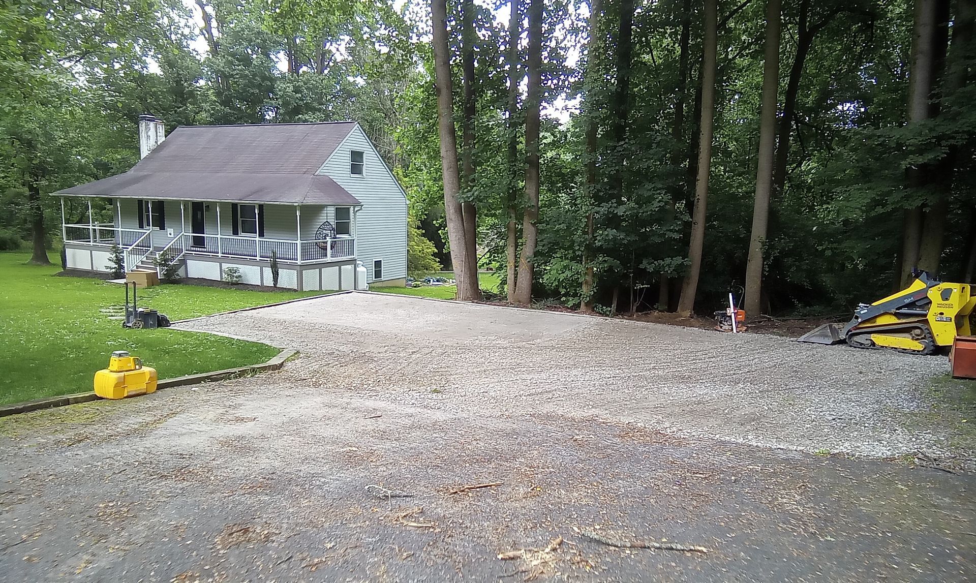 Gravel driveway beside a small white house in a wooded yard, with a yellow excavator at right.