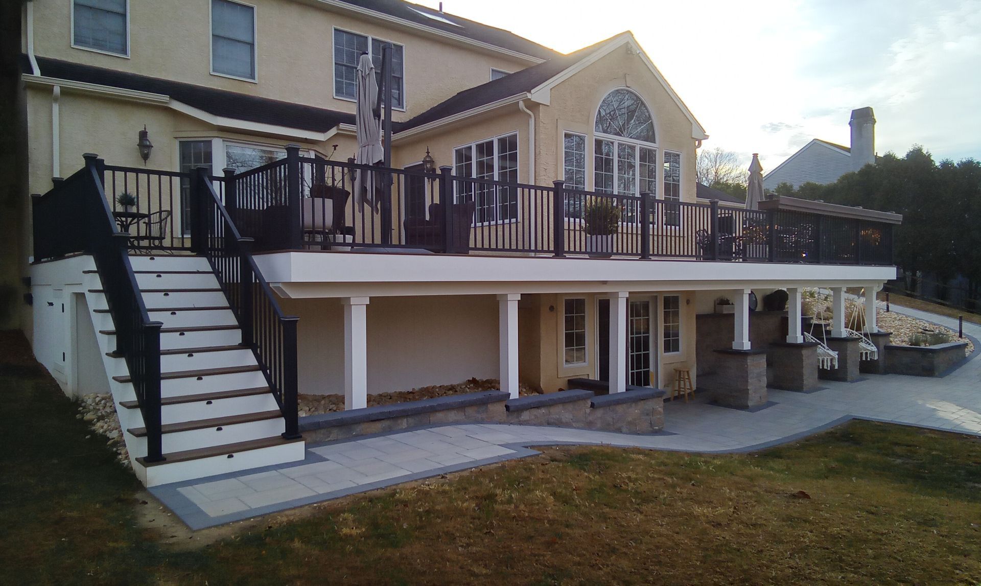 Two-story house with a white-railed deck, exterior stairs, and stone patio in a yard