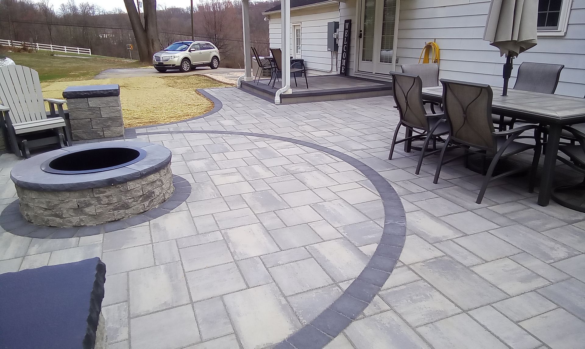 Patio with stone fire pit, curved pavers, outdoor dining table, and house siding in a backyard.