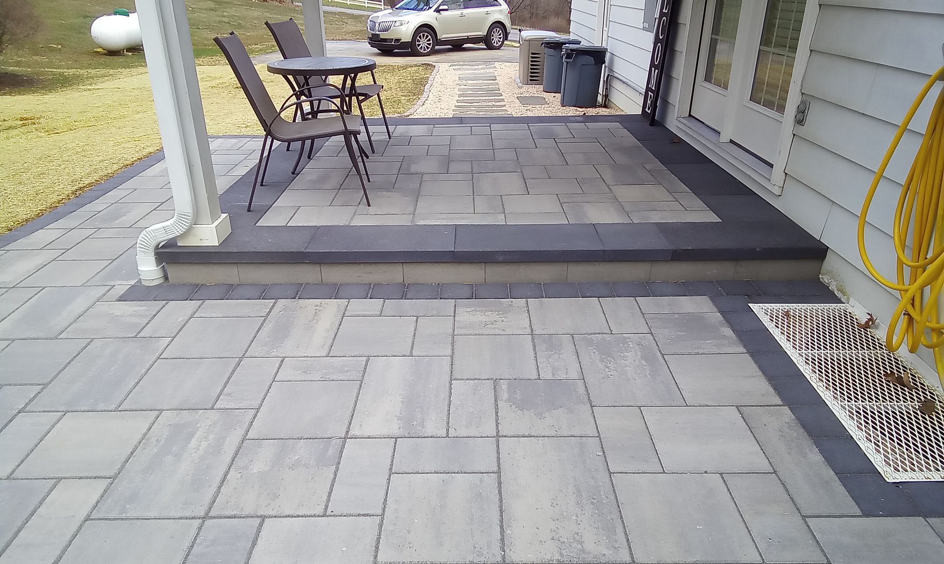 Patio with gray pavers, a small table and chairs, and steps leading down to a driveway with a parked car
