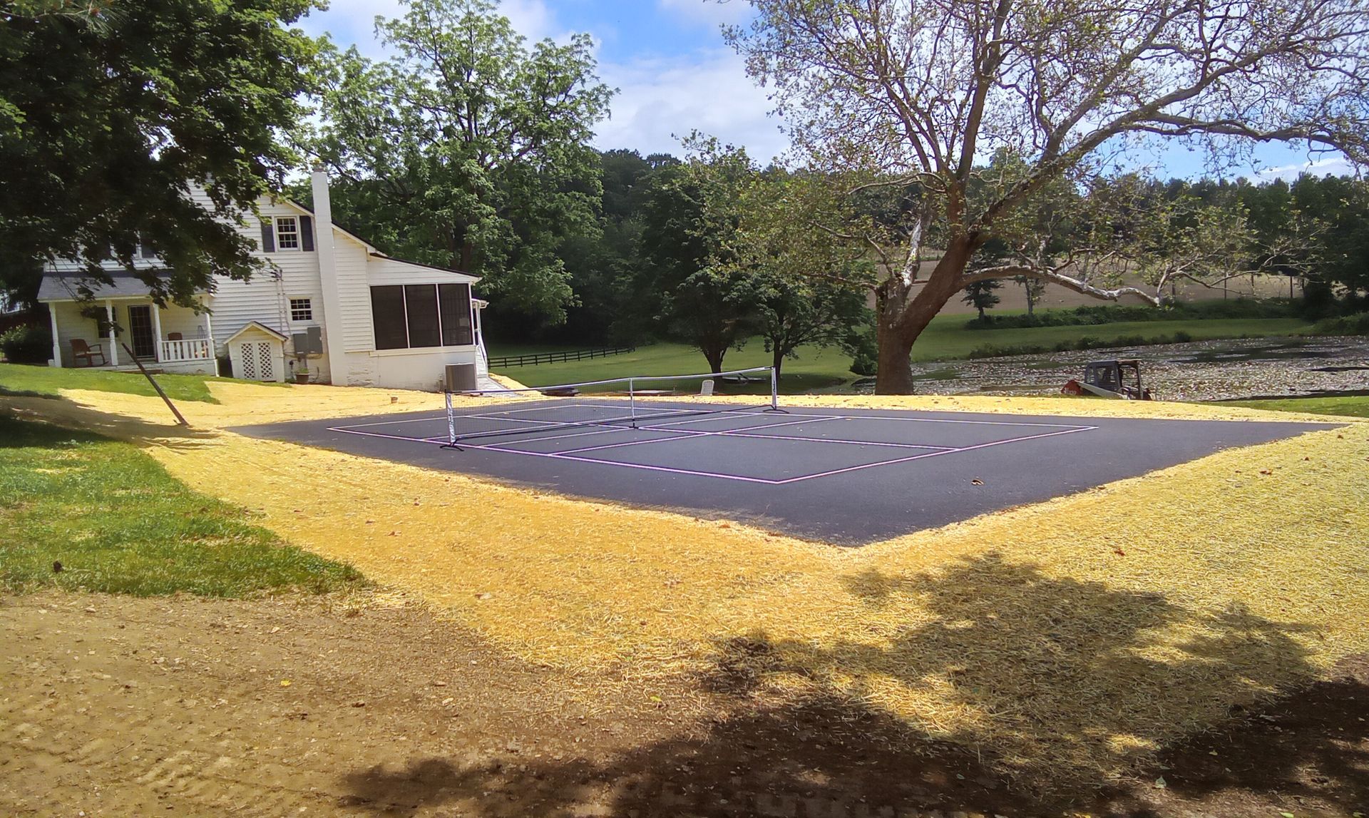 Outdoor volleyball court with black surface, yellow border, and surrounding trees near a house