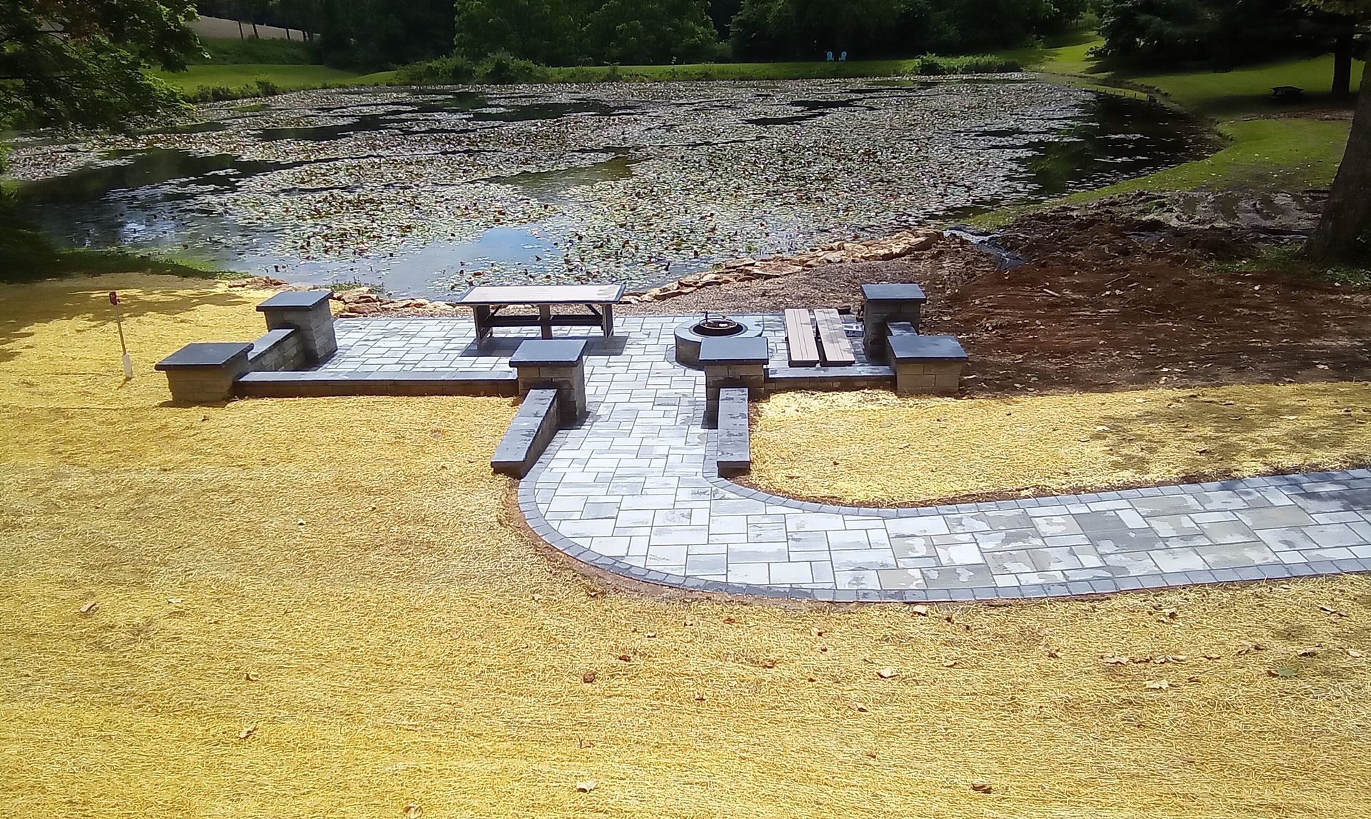 Stone walkway and benches beside a calm pond, bordered by yellow grass and trees.