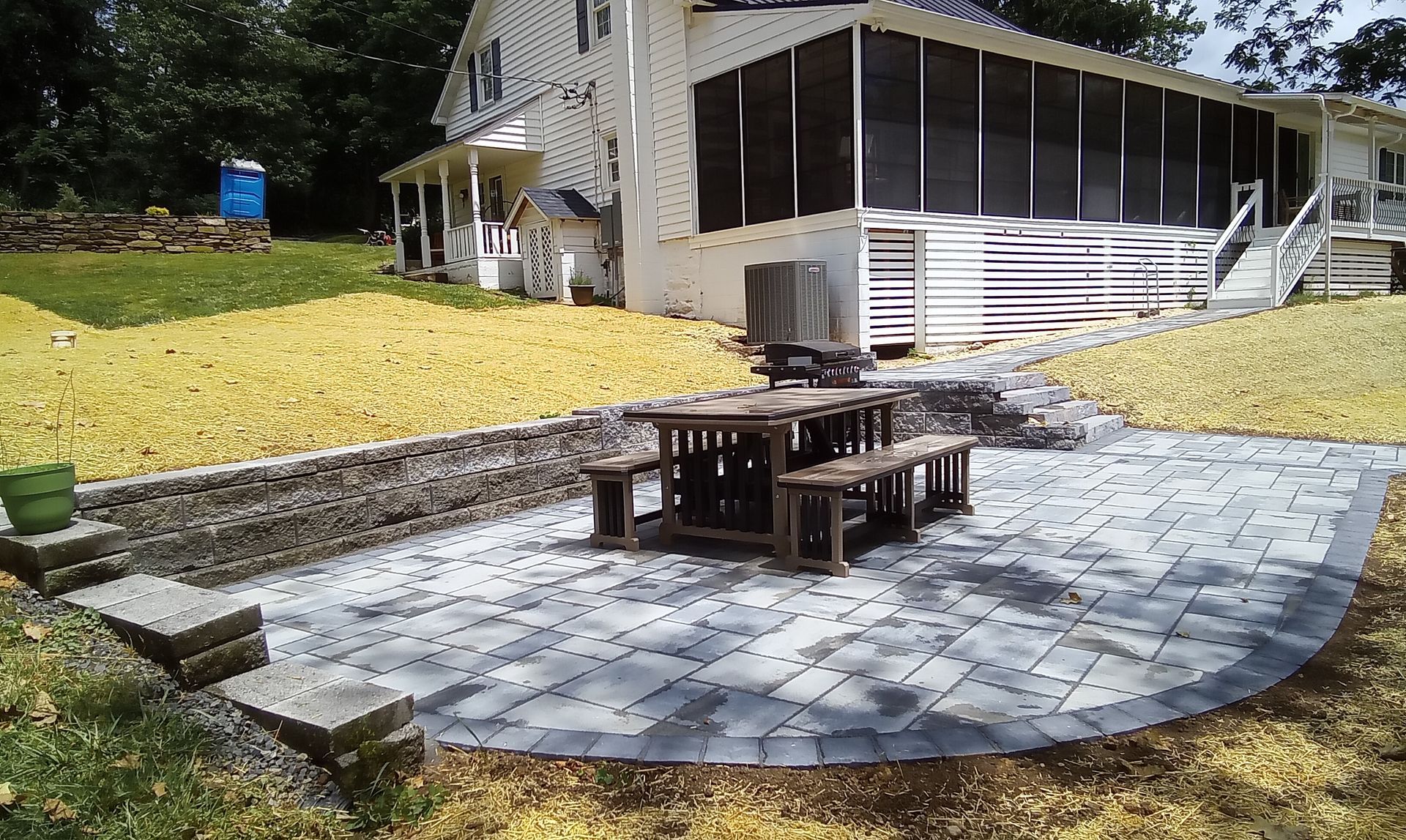 Backyard patio with stone pavers, a central fire pit table, and a house with screened porch and yellow ground cover.