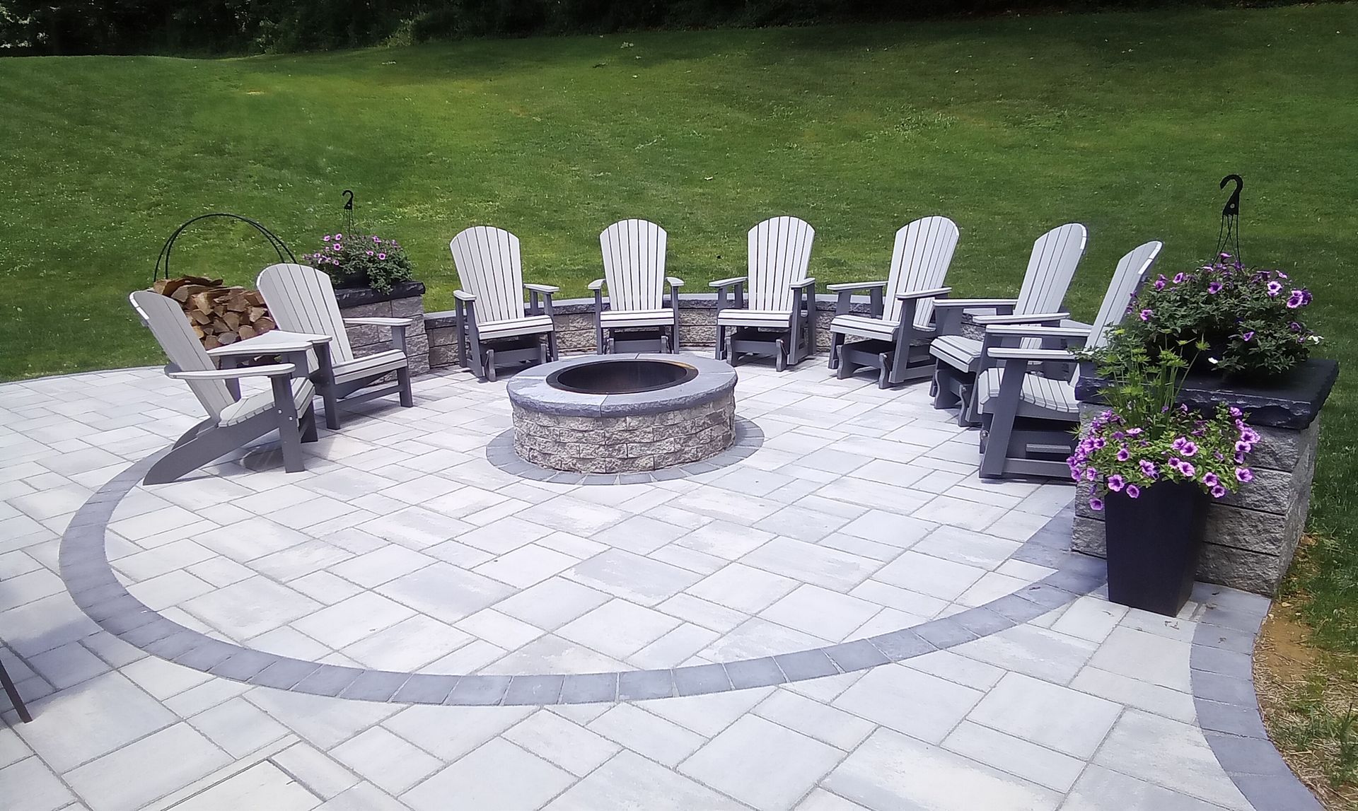Patio fire pit with white and black chairs arranged in a circle on a stone patio, beside flower planters.