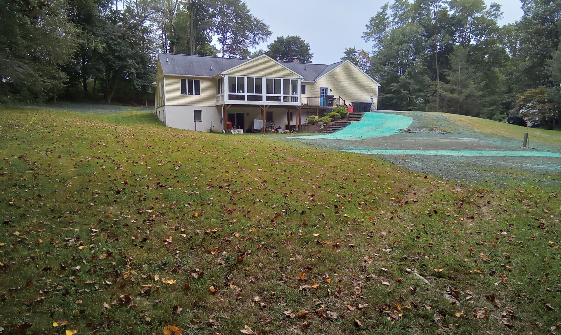 House on a grassy hillside with a deck and blue tarp, surrounded by trees