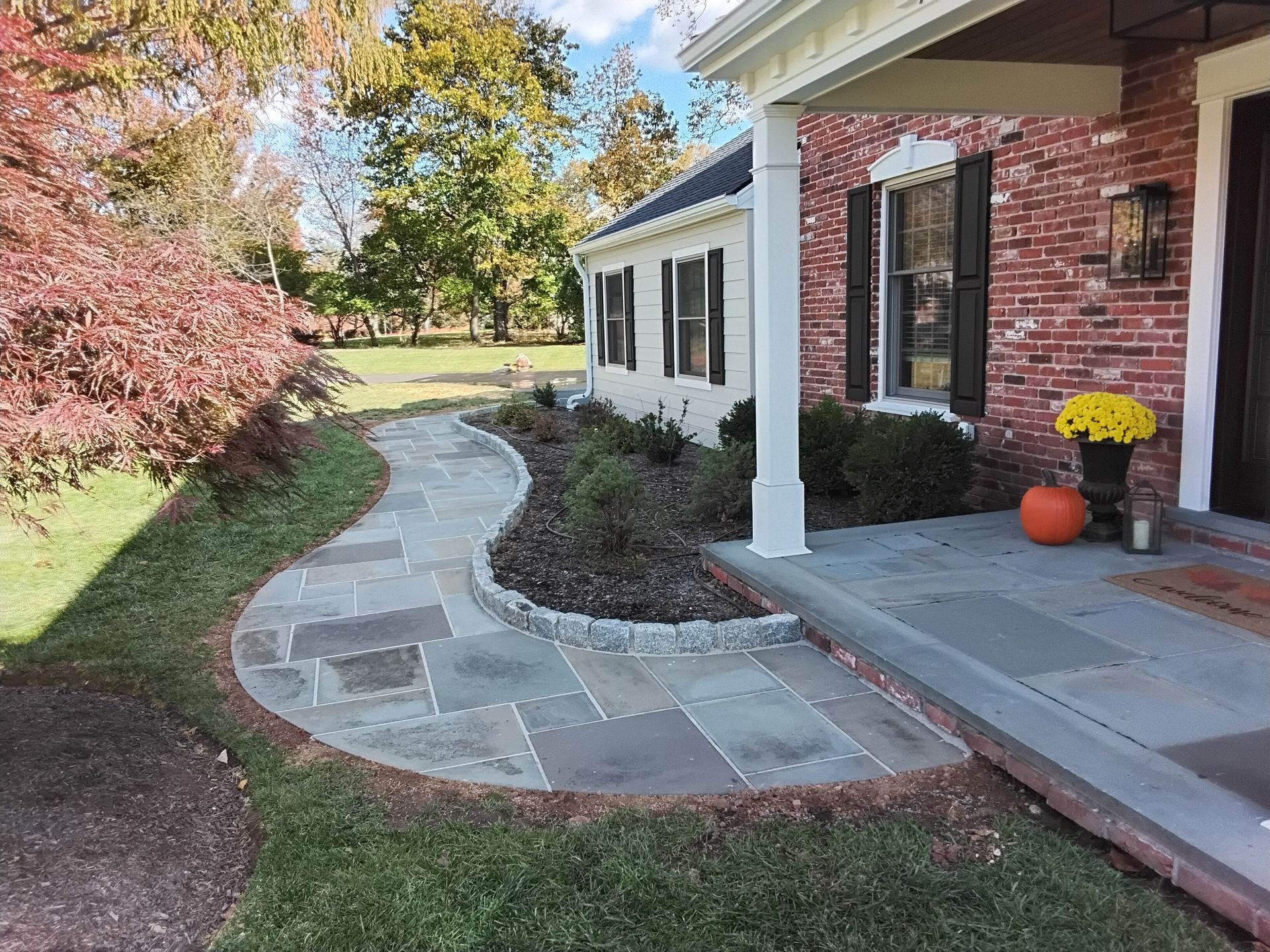 Curved stone walkway beside a red brick house, with porch columns, fall foliage, and pumpkins by the door