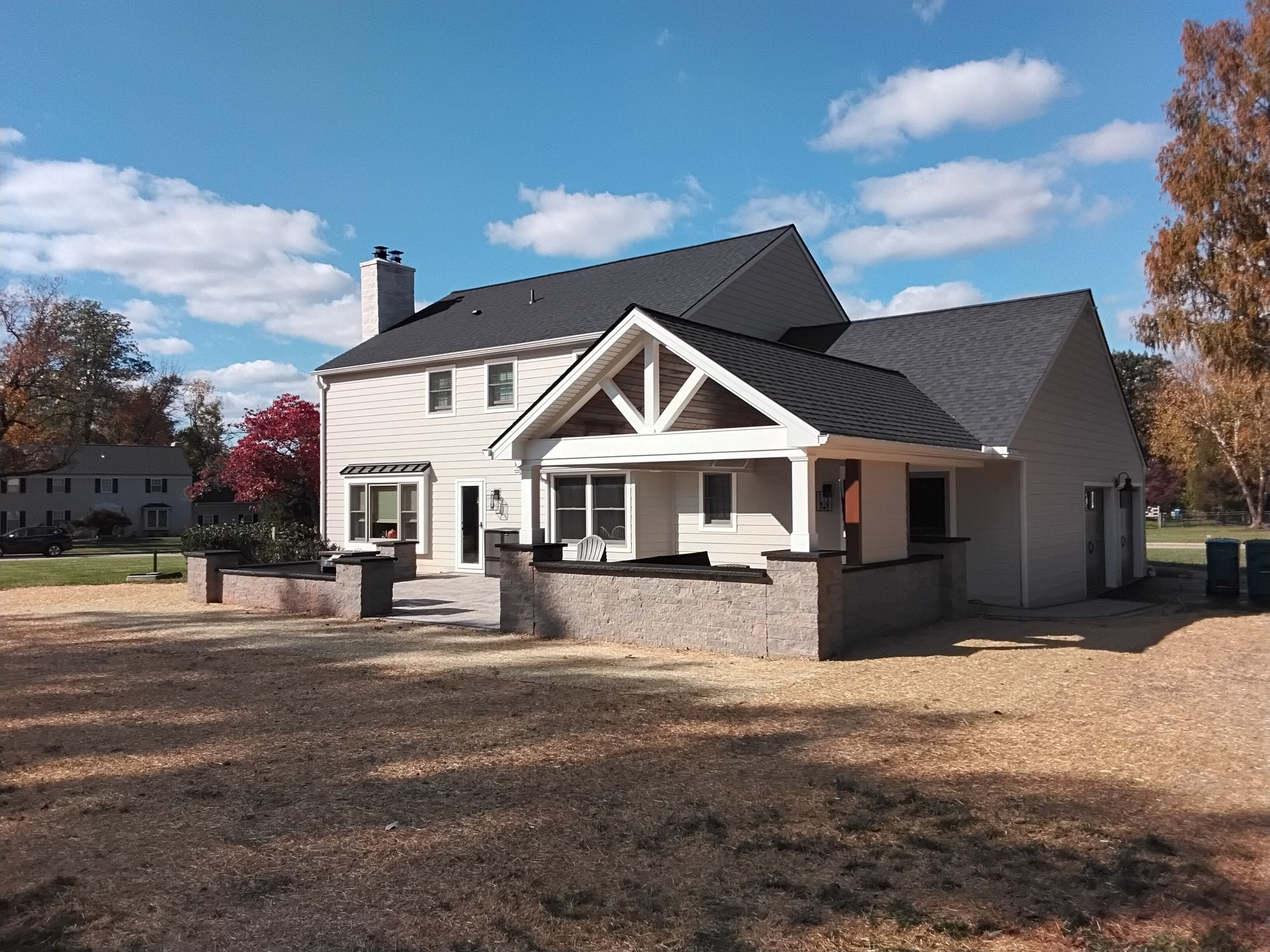 White house with dark roof and front porch on a sunny day, surrounded by a gravel yard and trees.