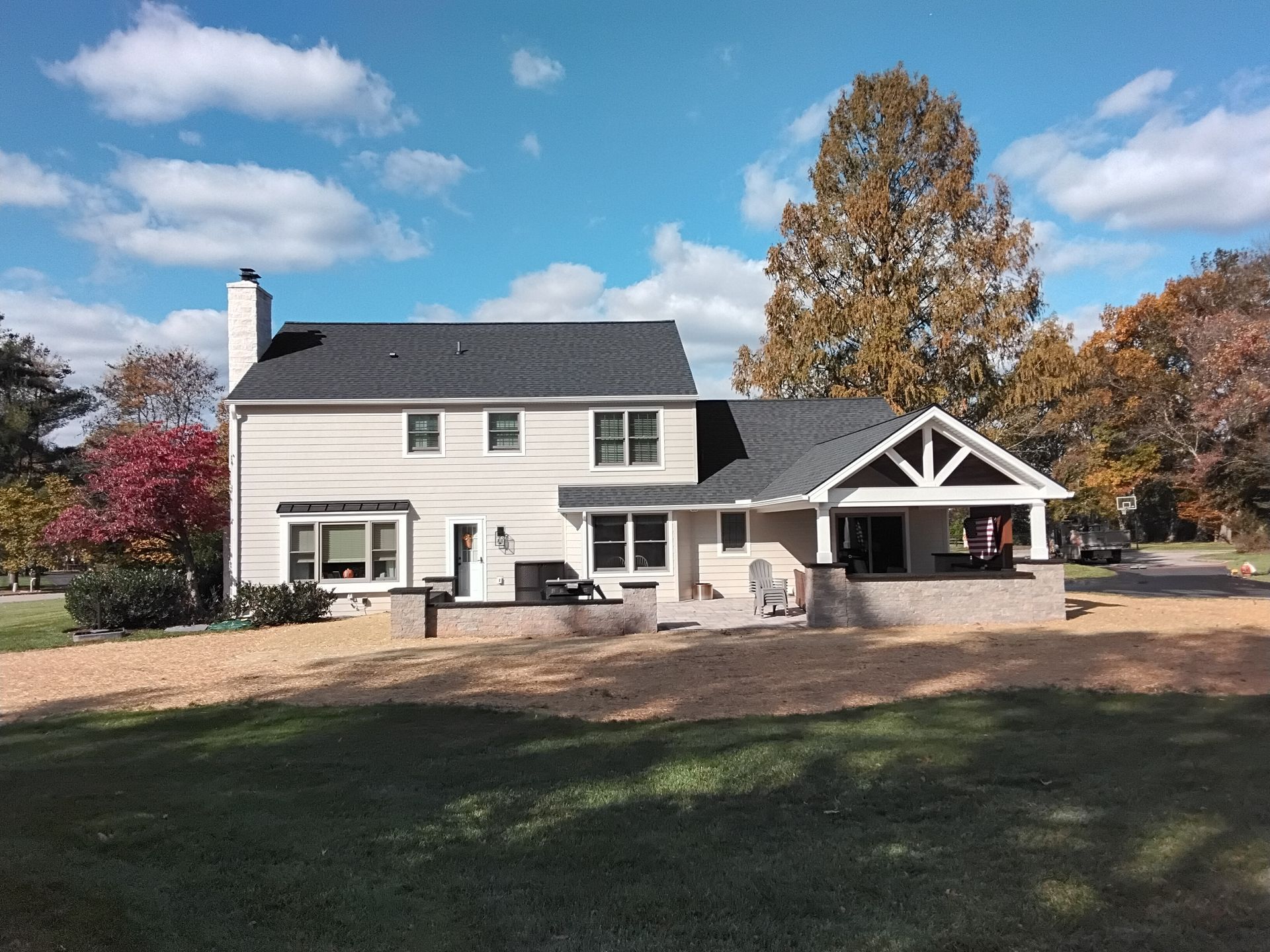 White two-story house with black roof, porch, and autumn trees under a blue sky