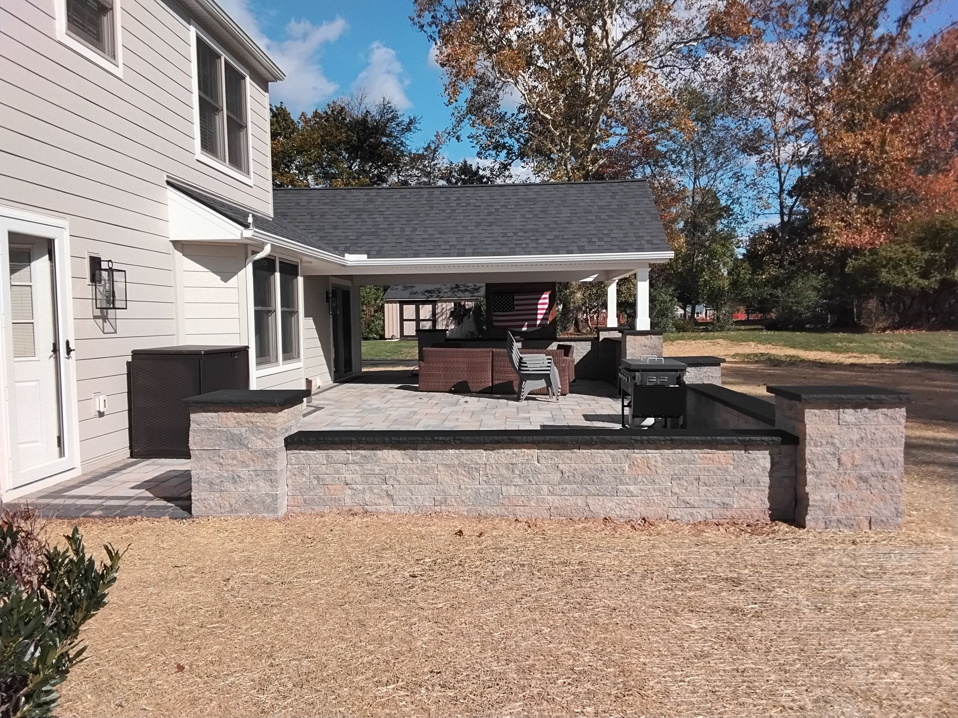 House patio with covered deck, brick seating wall, grill, and fall trees in the yard