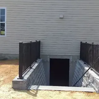Exterior basement door with stairs. Black door and railings. Gray retaining walls and siding.