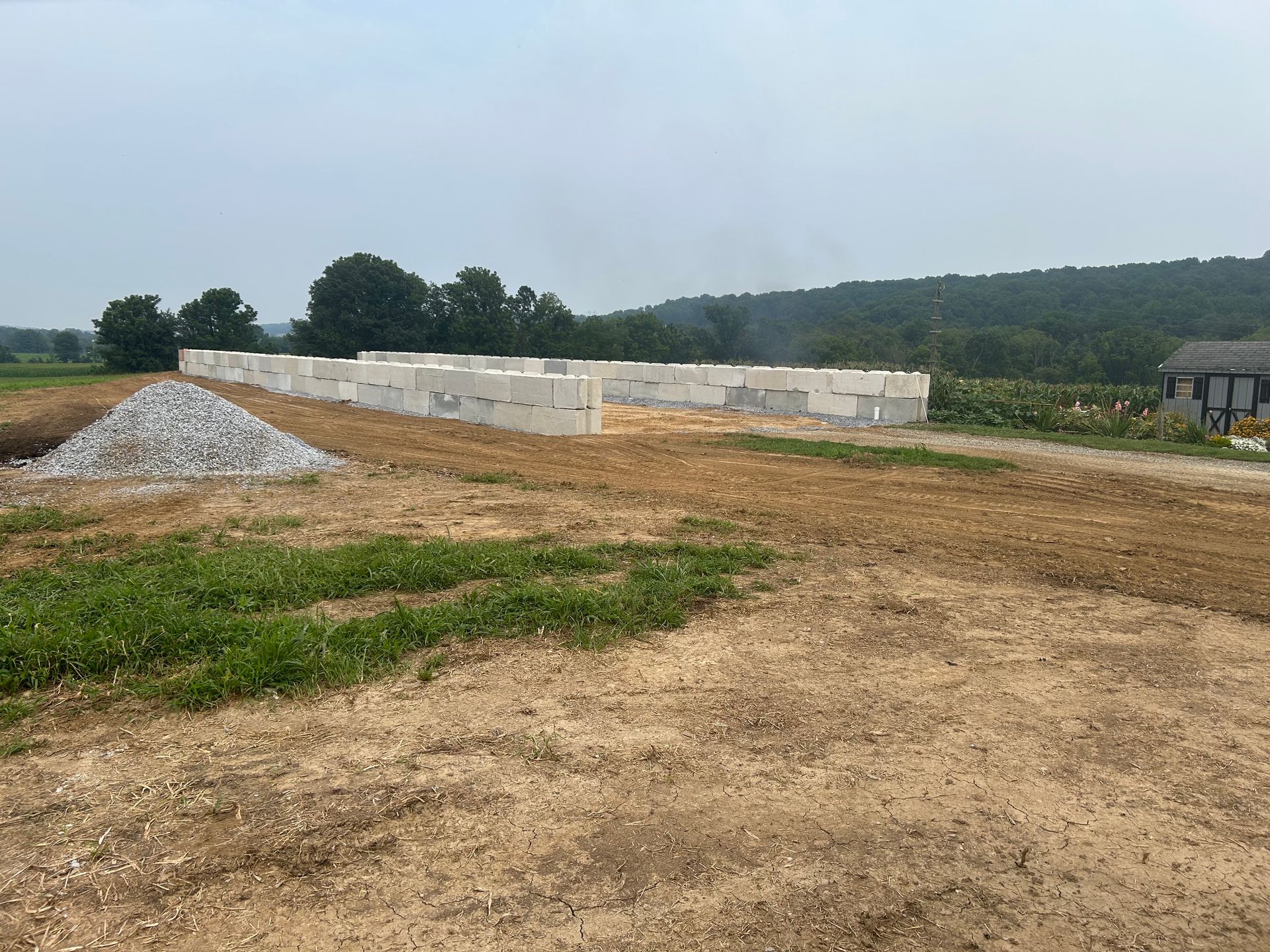 Construction site with concrete block foundation, pile of gravel, and farmland in the background.