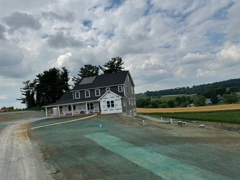 Rural farmhouse by a gravel road under cloudy skies, with open fields and a sloping driveway