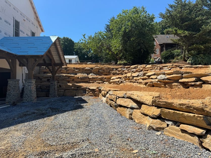 Stone retaining walls with gravel path leading to a wooden shelter, blue sky, and trees.