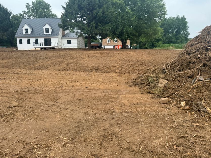 Cleared dirt lot beside a white house with trees and a woodpile on the right