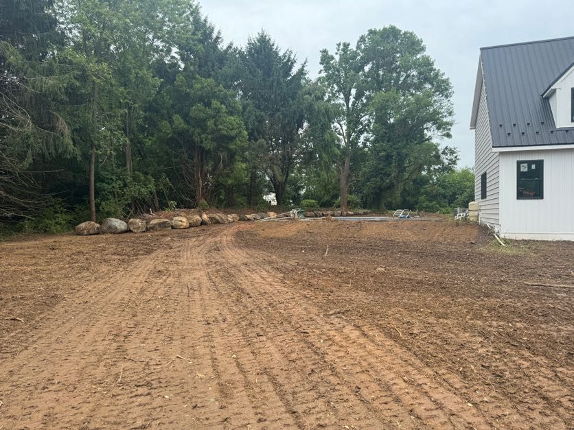 Freshly graded dirt lot beside a white house, with trees and rocks along the edge.