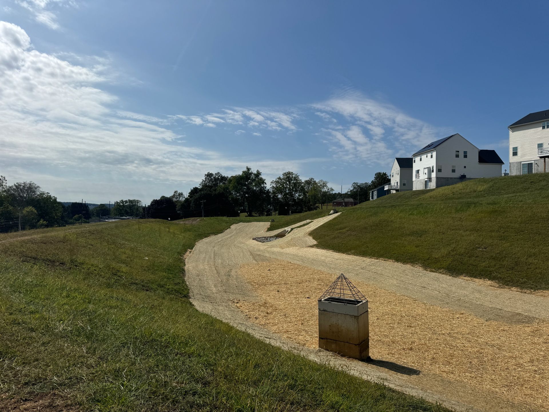 Winding gravel path through a grassy hill toward white houses under a blue, cloudy sky