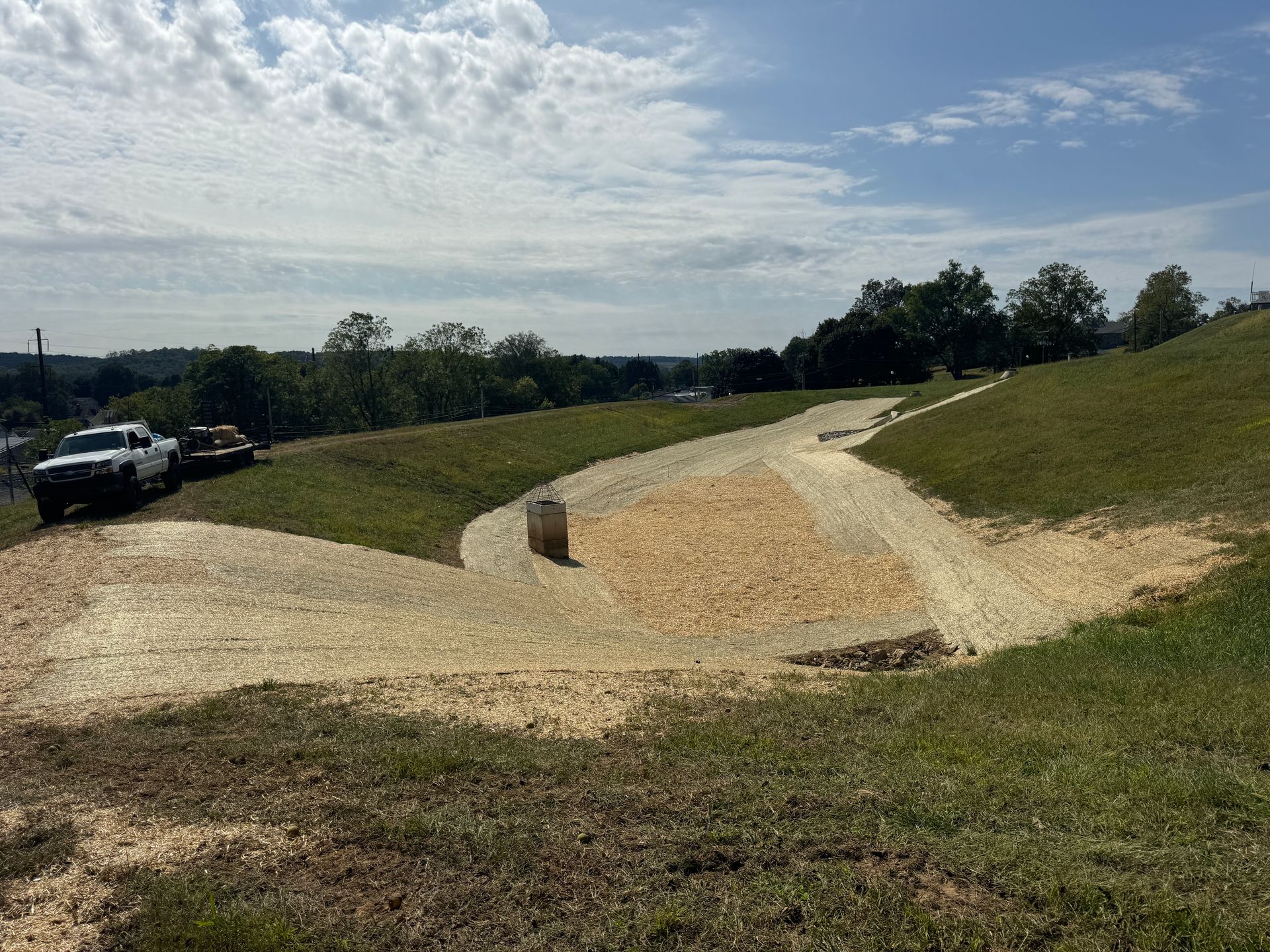 Gravel path winding through a grassy park under a cloudy sky, with parked cars on the left.