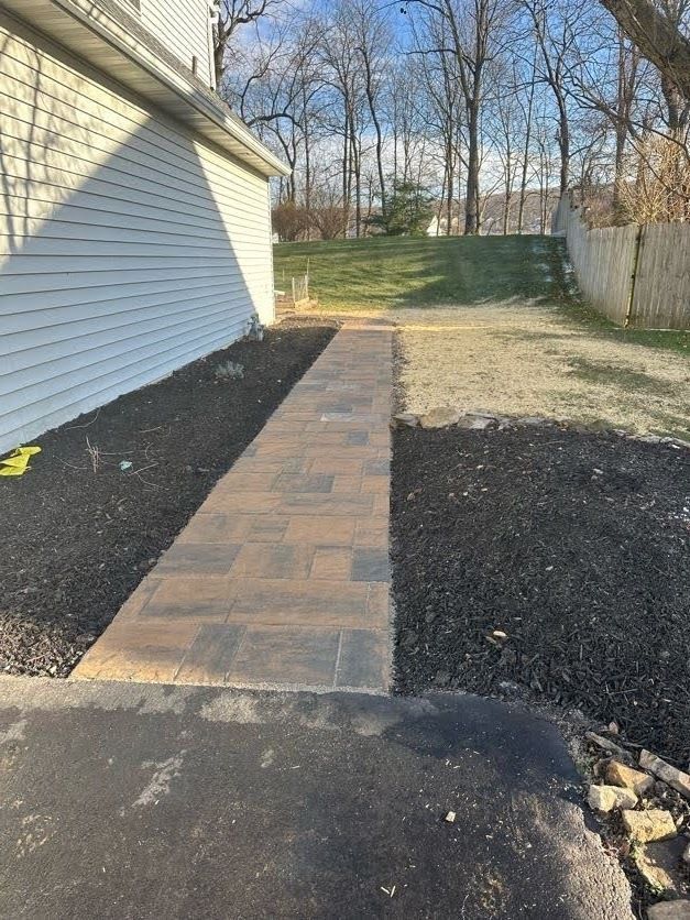 Brick walkway beside house leading to grassy yard and fenced edge in a sunny backyard