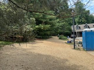 Gravel driveway beside trees and a house, with a blue portable toilet on the right.