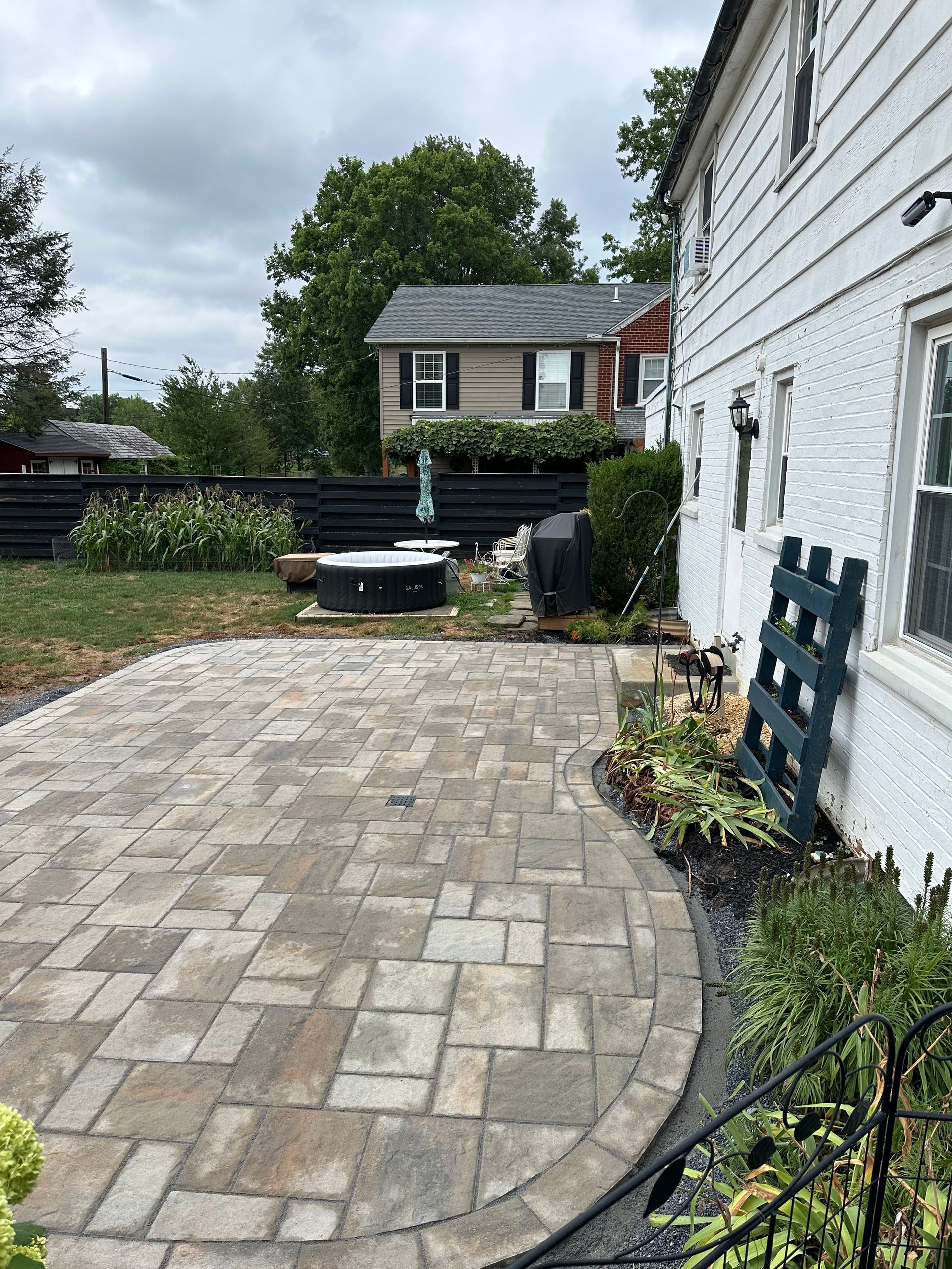Stone patio with curved pavers beside a white house, leading to a backyard and neighboring homes.