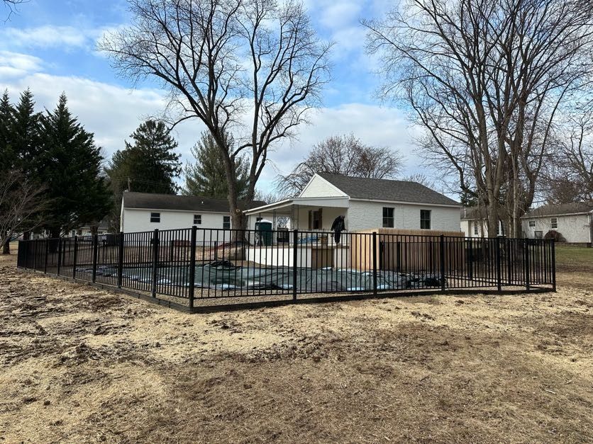 Backyard pool enclosed by black fence beside a white house and leafless trees under a cloudy sky