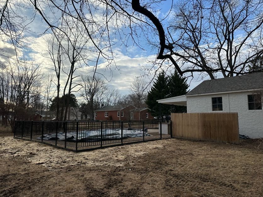 Backyard with fenced pool, leafless trees, and a white house under a cloudy sky