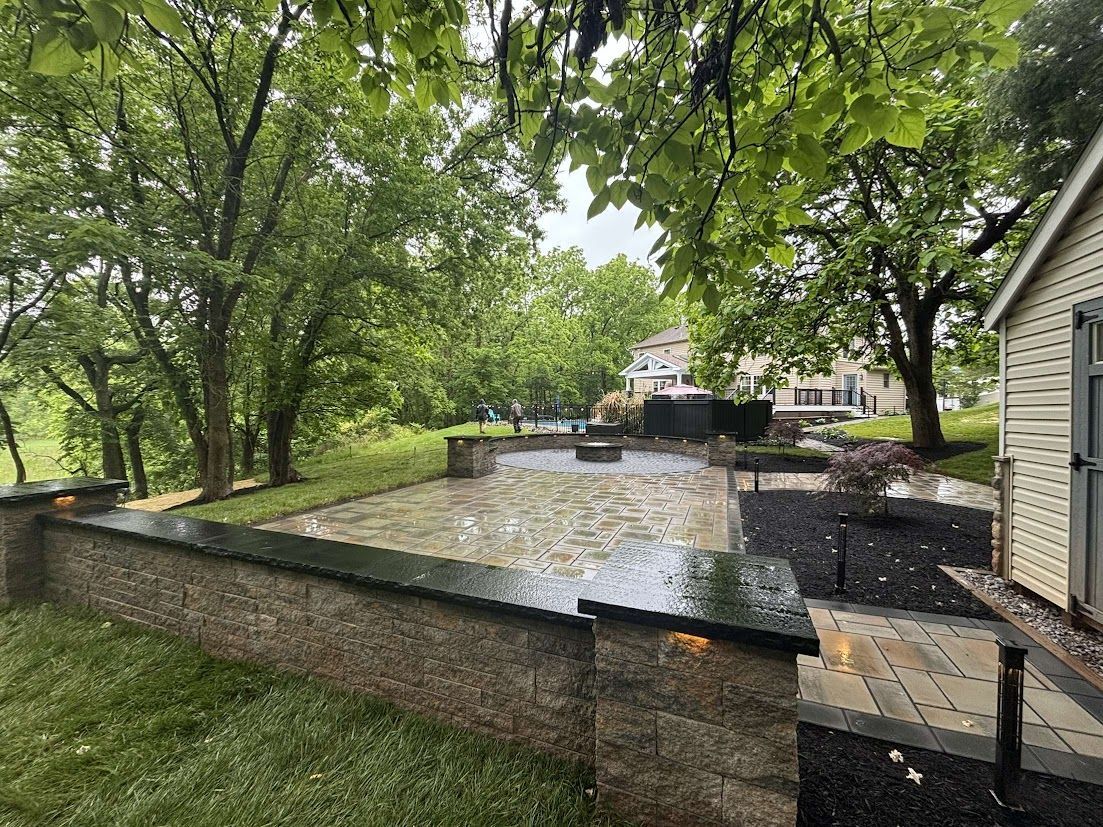 Stone patio with low walls and a building in a green, tree-filled outdoor space.