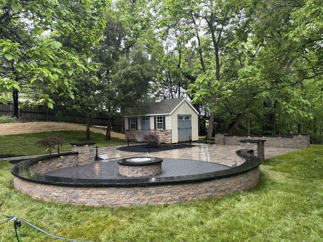 Circular stone patio with fire pit, small shed in the background, surrounded by trees and grass.