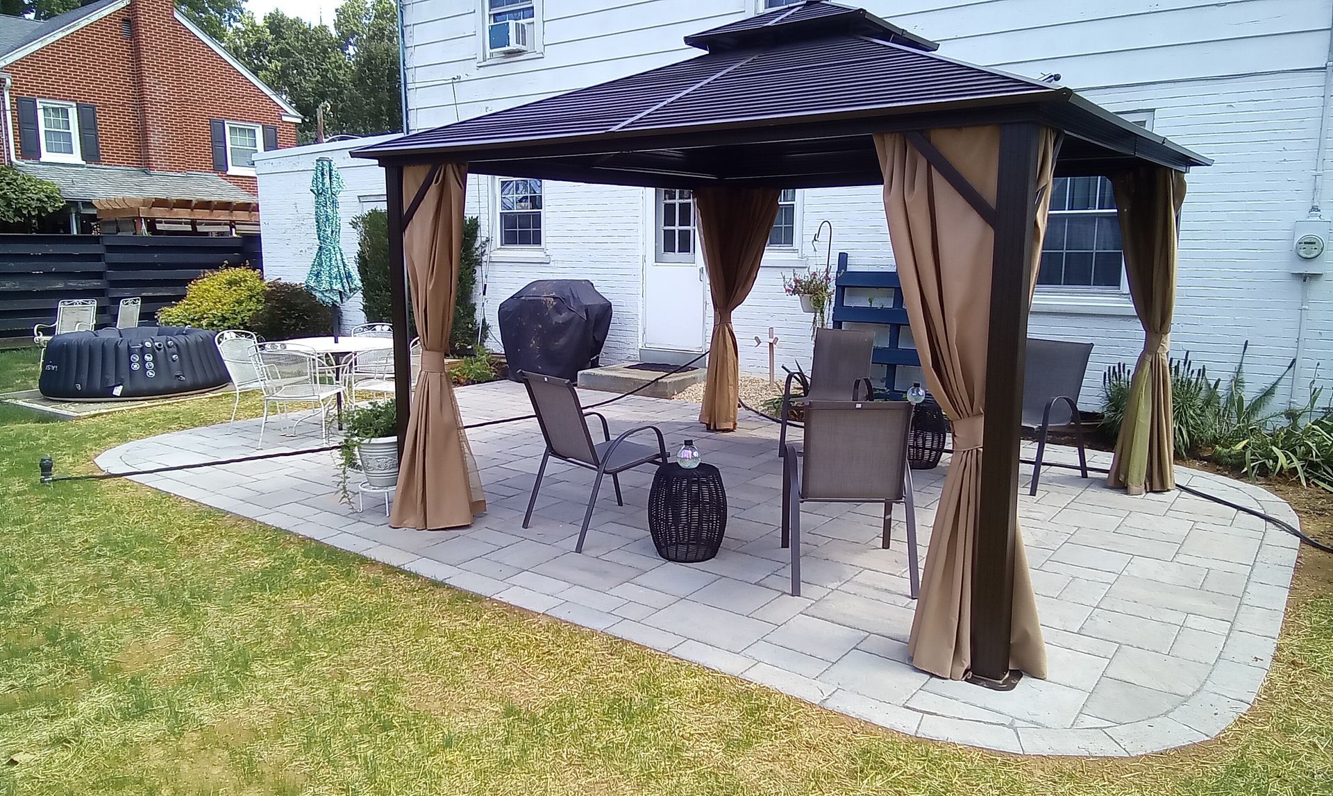 Gazebo on a brick patio with outdoor furniture, brown curtains, and a grill in a backyard.