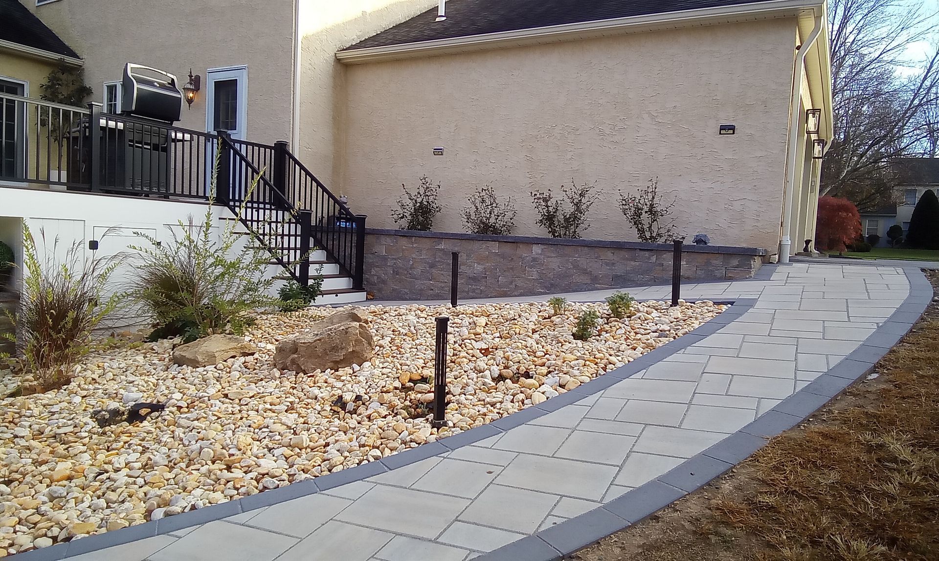 A paved walkway curves through a landscaped yard with a retaining wall, leading to a house.
