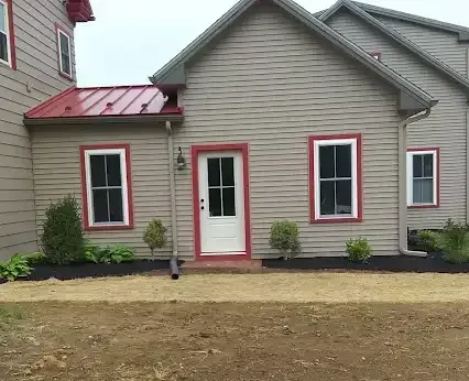 Tan house with red-trimmed white windows and door, red metal roof, and black mulch landscaping.