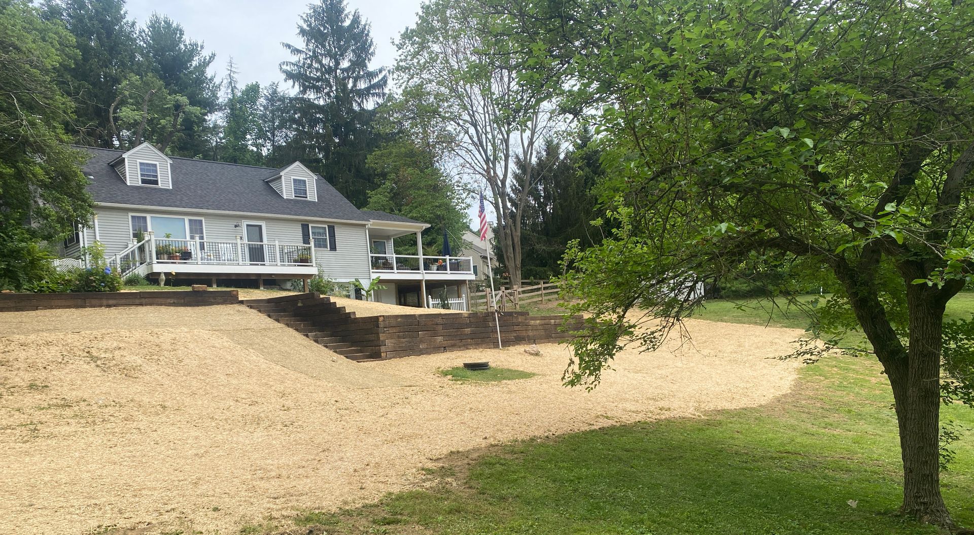 White house with black roof, deck, and sloping yard covered in beige material. Trees surround the house.