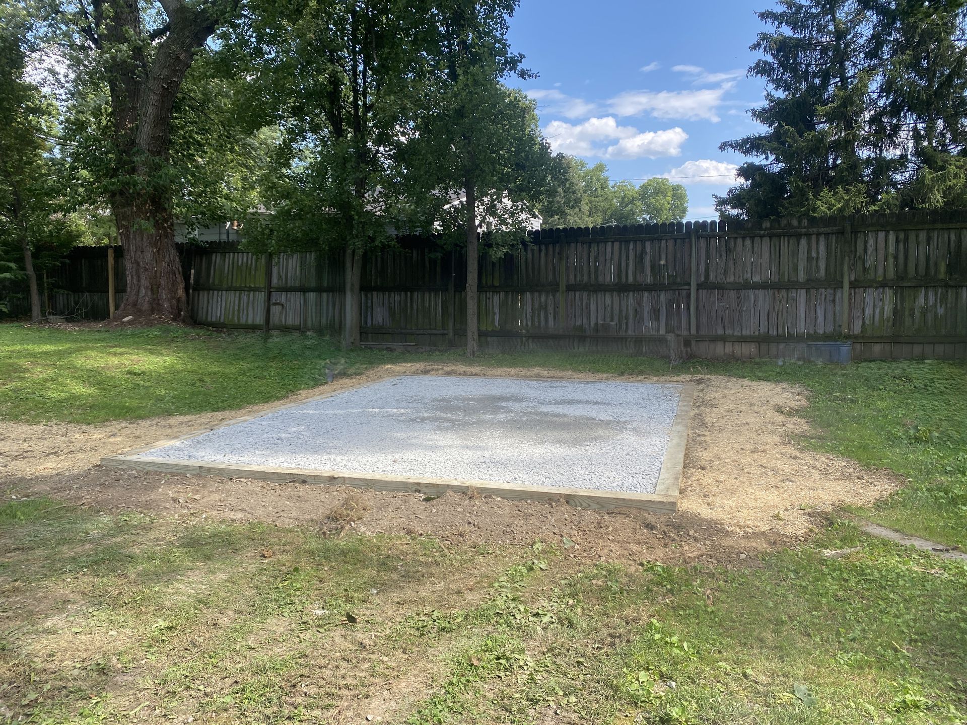 Gravel pad in backyard, bordered by dirt. Brown fence and green trees in the background. Sunny day.