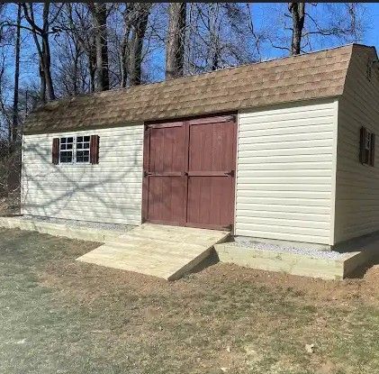 Tan shed with brown roof and doors, wooden ramp in front, set in a grassy yard.