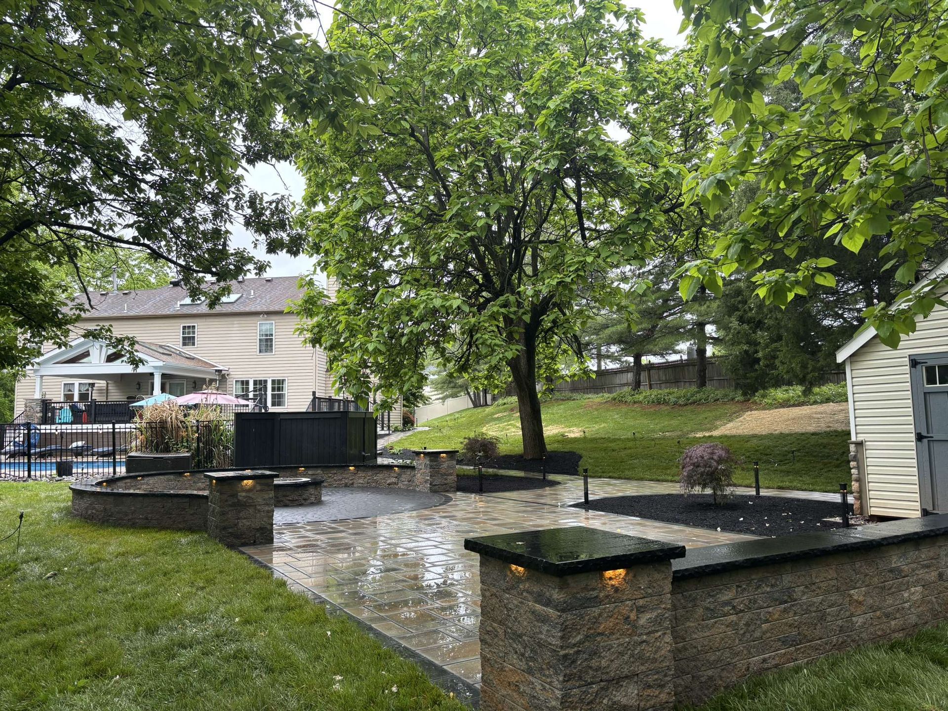 Rainy backyard patio with stone walls, lit pillars, trees, and a house in the background
