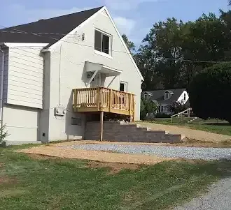 Two-story house with wooden deck and steps. Gravel and brick retaining wall. Green grass and trees in background.