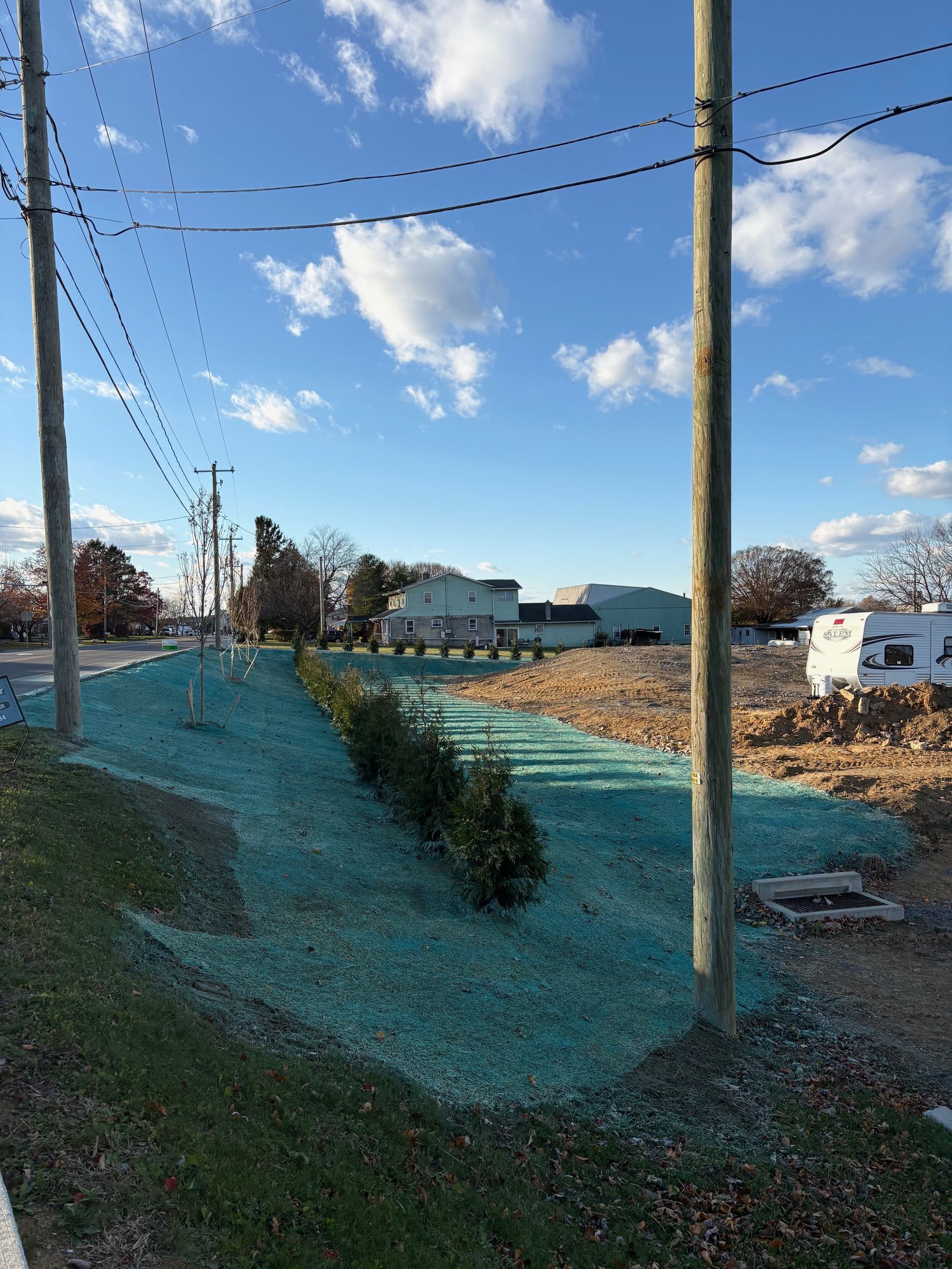 Rural roadside with utility poles, wires, a blue sky, and a house in the distance amid grassy fields.