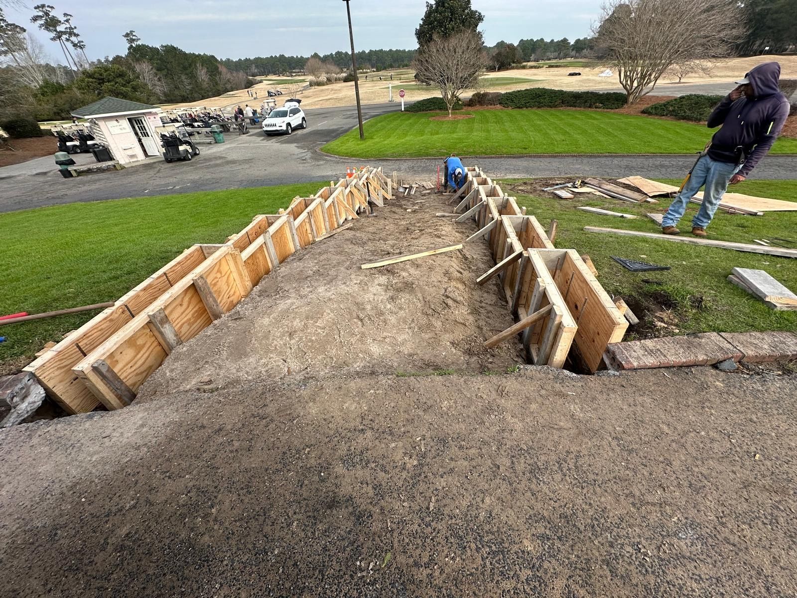Construction site with wooden forms for a long concrete path, two workers, and a gravel slope