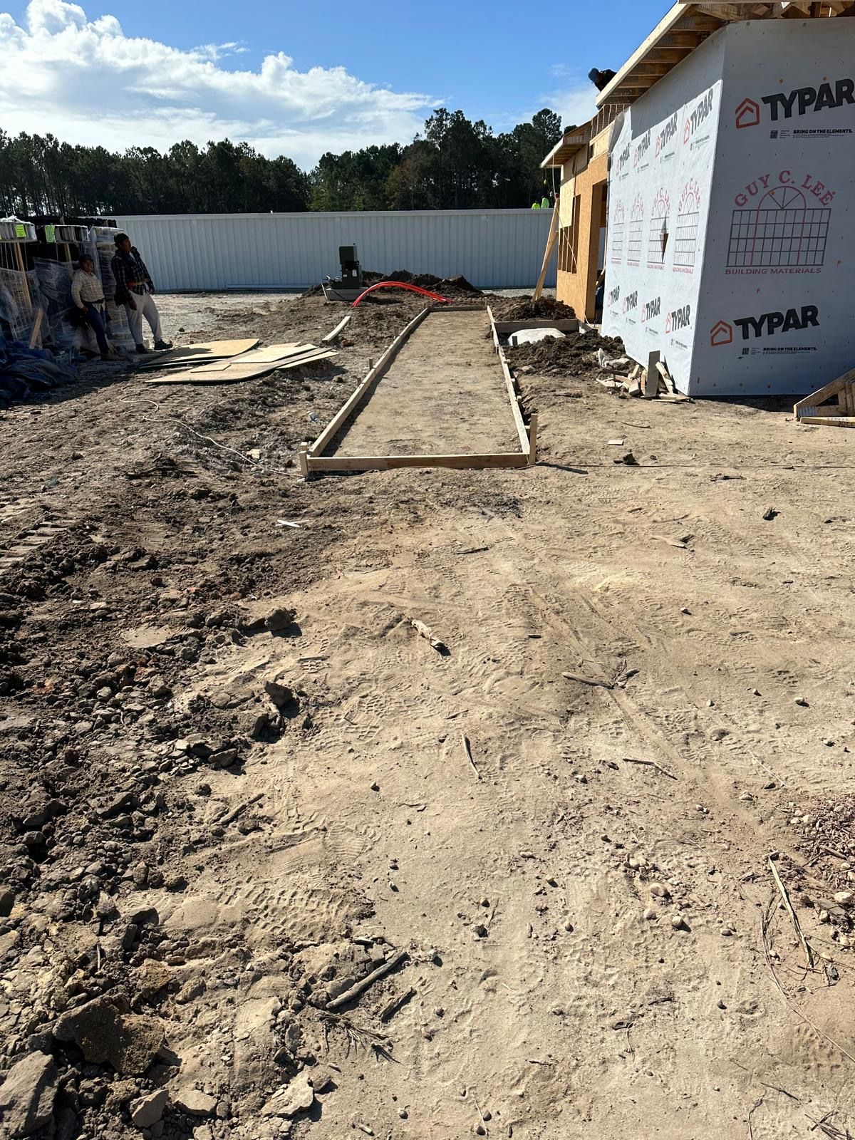 Construction site with dirt path and timber forms beside a house wrapped in Typar insulation.
