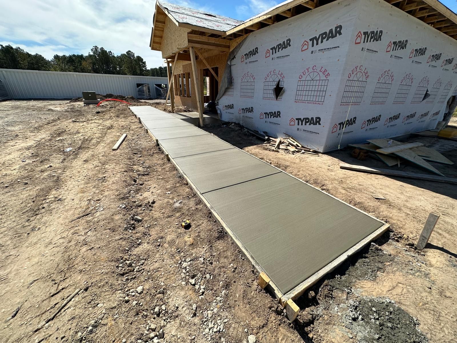 Concrete walkway leading to a house under construction with wrapped exterior walls and exposed framing